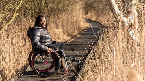 Ade Adepitan sat in his wheelchair, smiling, on the boardwalk at Wicken Fen